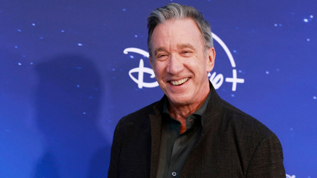 Tim Allen smiles in front of a blue background with "Disney" printed on it. 