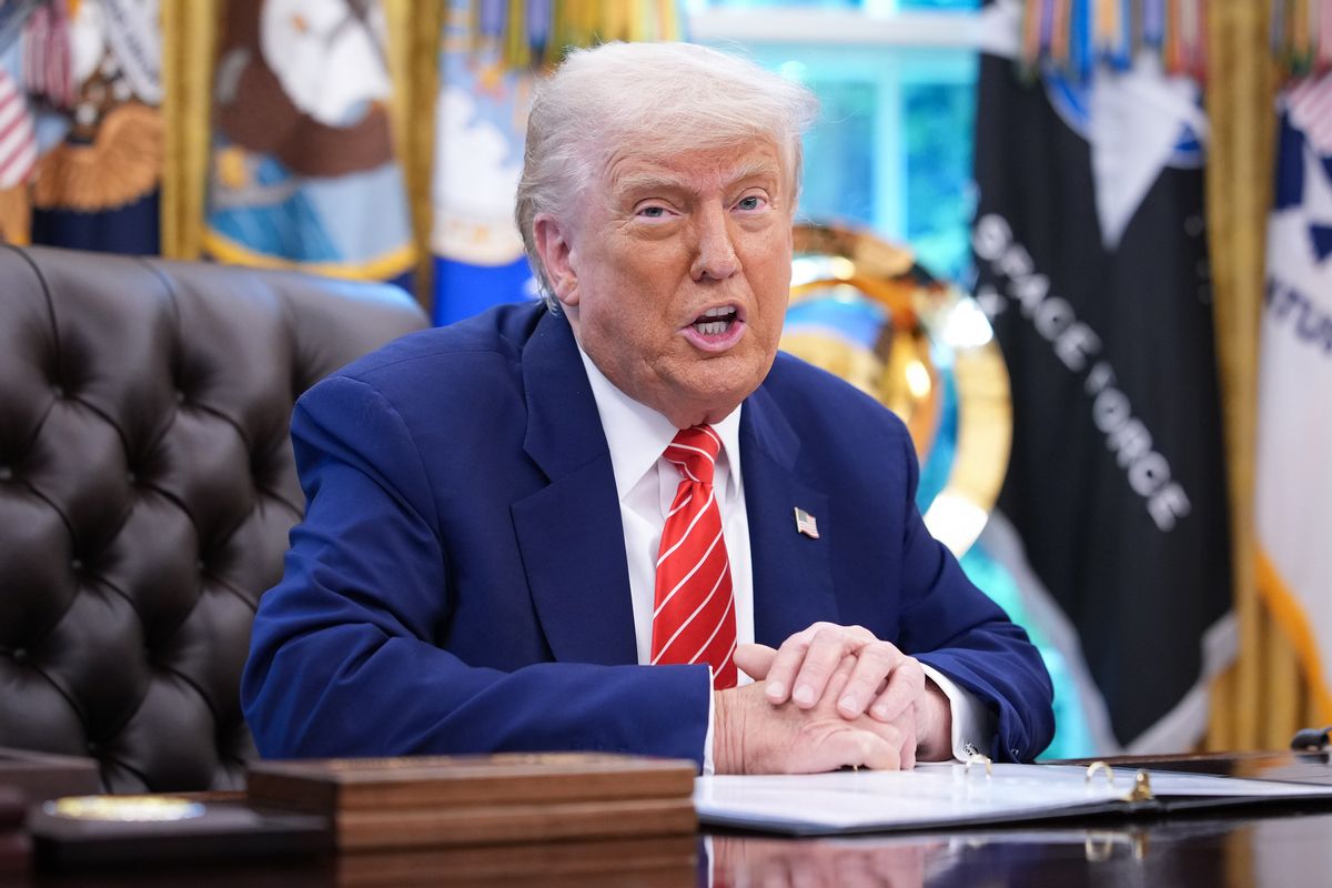A white man wearing a blue suit speaks while sitting at a desk. His hands are clasped, and a binder of documents appears to be in front of him.