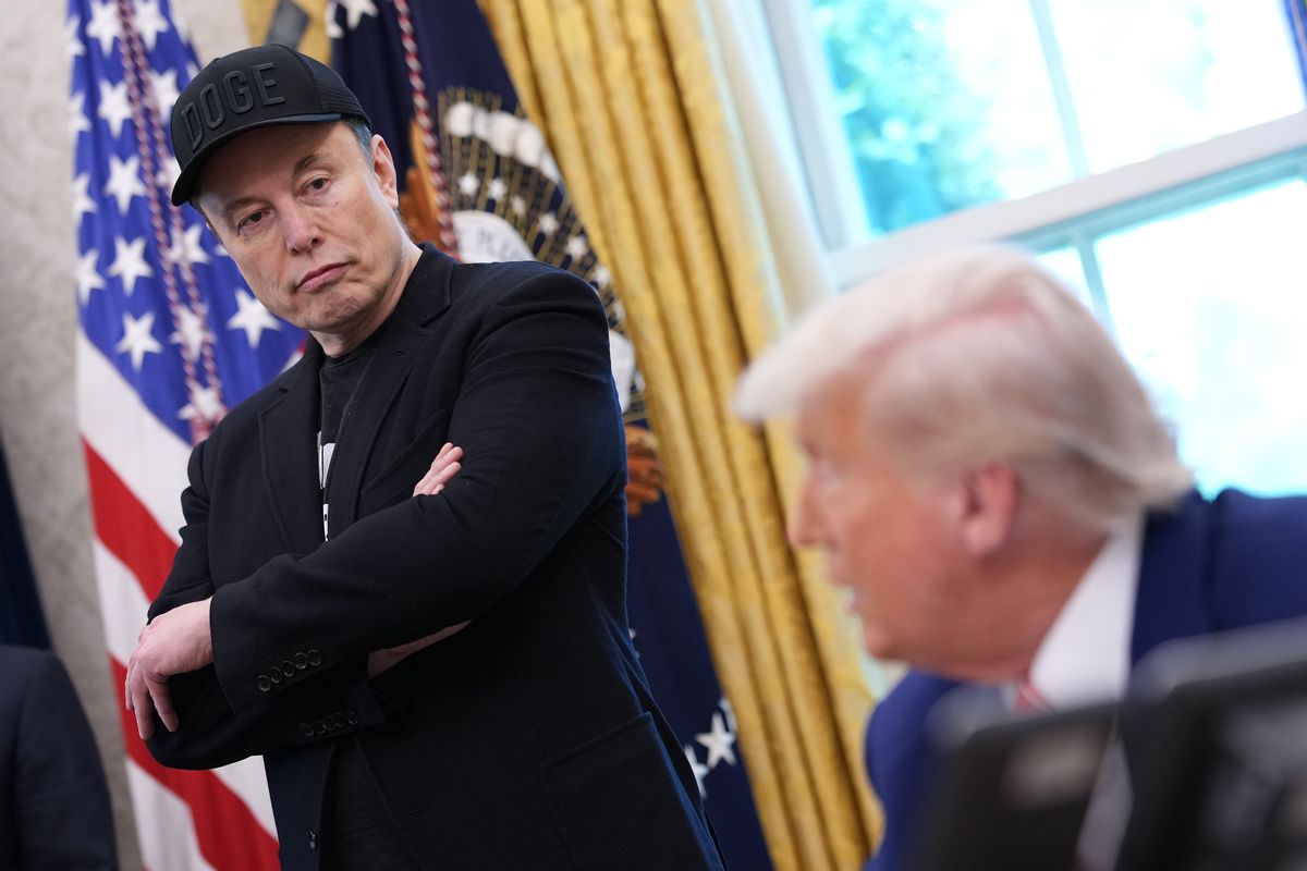 A white man wearing all black that's standing up looks down at a white man wearing a blue suit. The man wearing black is the focus of the photo.