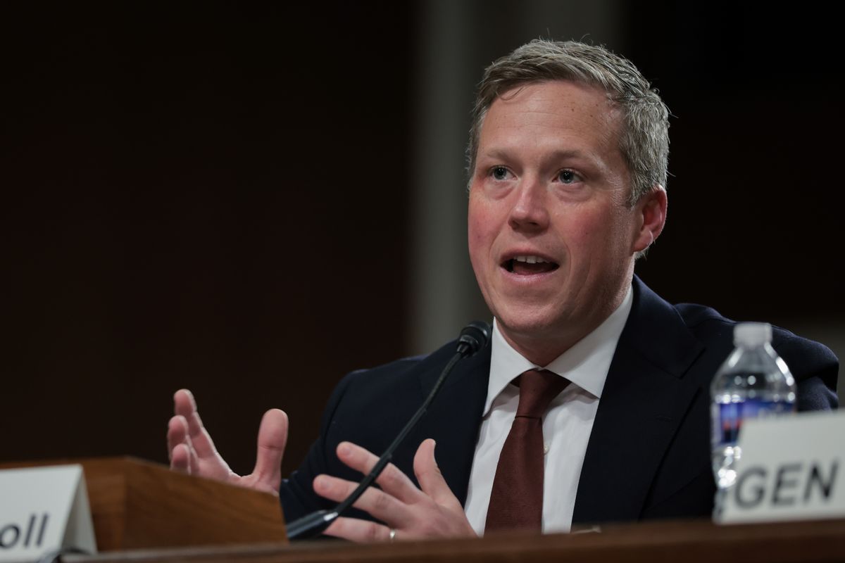 A white man speaks behind a microphone while sitting down.