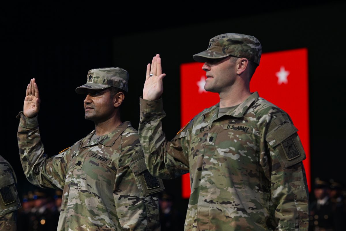 A South Asian man and a white man in camouflage military uniforms hold their palms up and appear to be swearing an oath. 