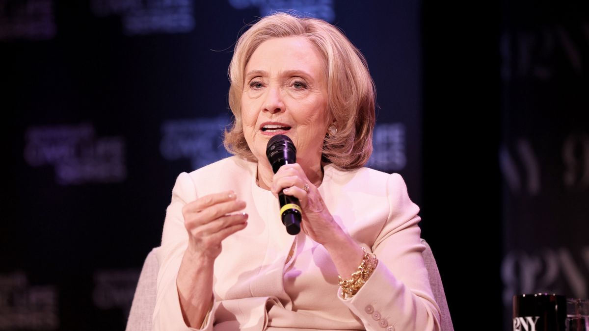 Hillary Clinton, wearing a light-colored blazer and gold bracelet, speaks into a handheld microphone during a public event. She is seated on stage in front of a dark backdrop with blurred white text.