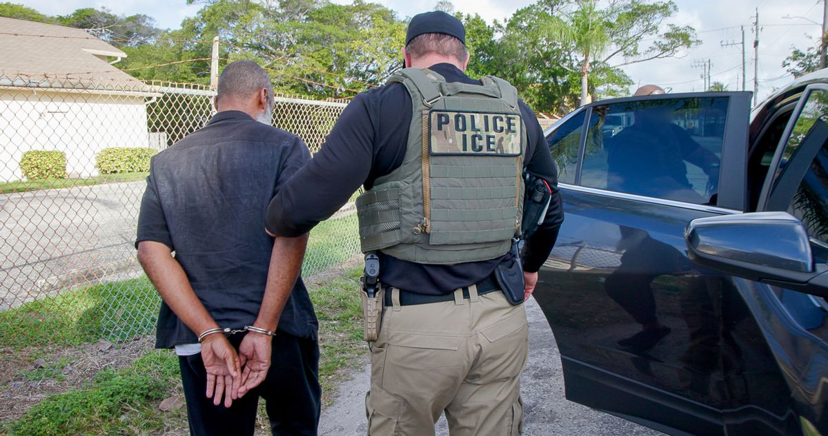 An ICE officer in a uniform holding the arm of a handcuffed person. 