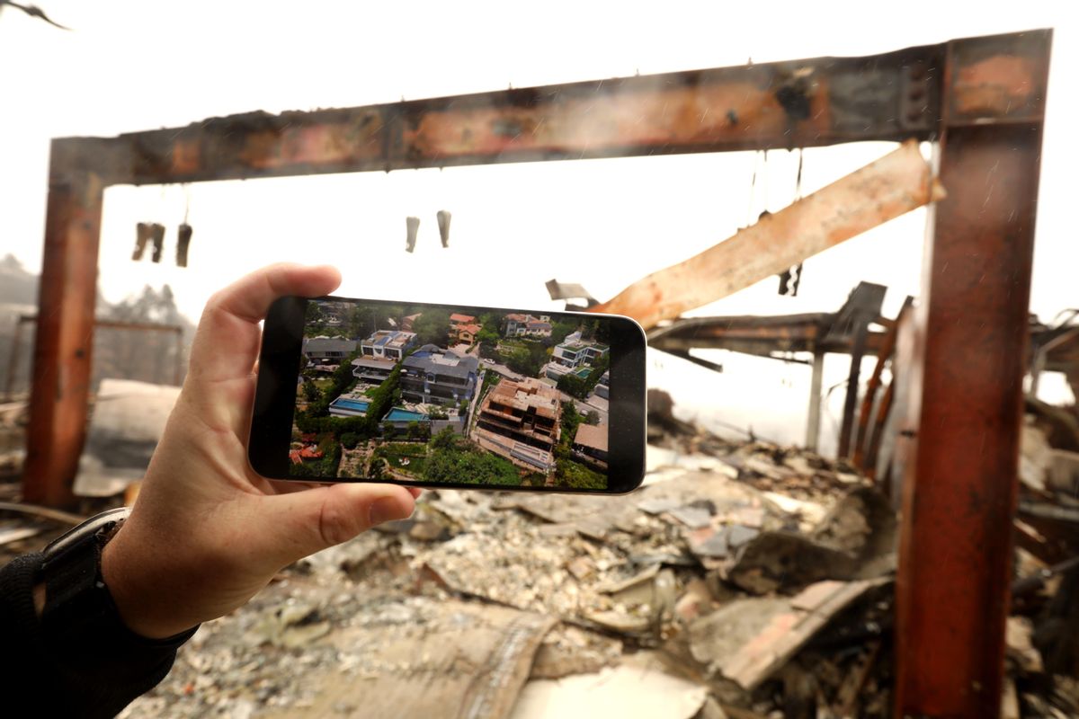 A hand holding a phone showing a large home. Behind the hand and phone is the ruins of that home after a fire. 