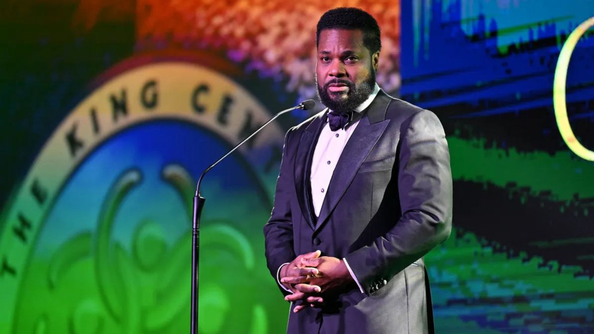 A Black man wearing a dark suit stands in front of a microphone.