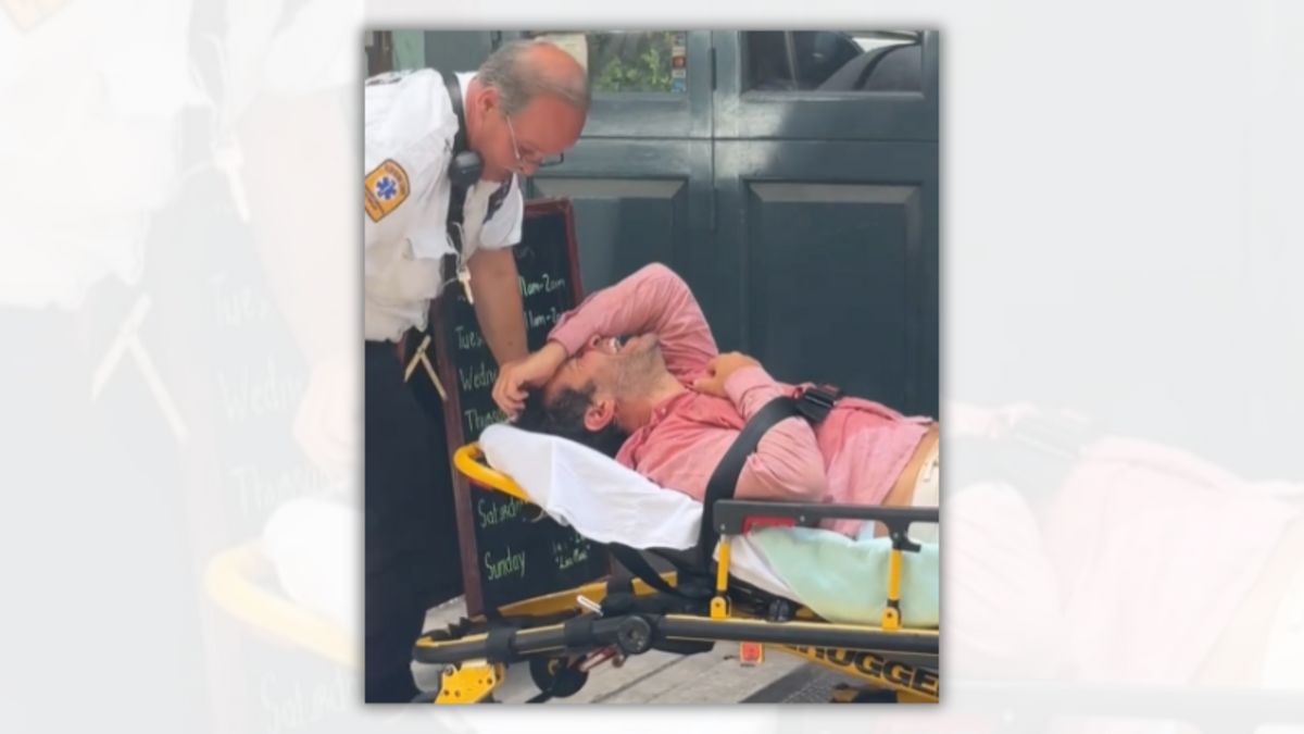 A white man wearing a pink shirt that looks wet has a hand over his face as he lies on a stretcher. A person looks down at him.