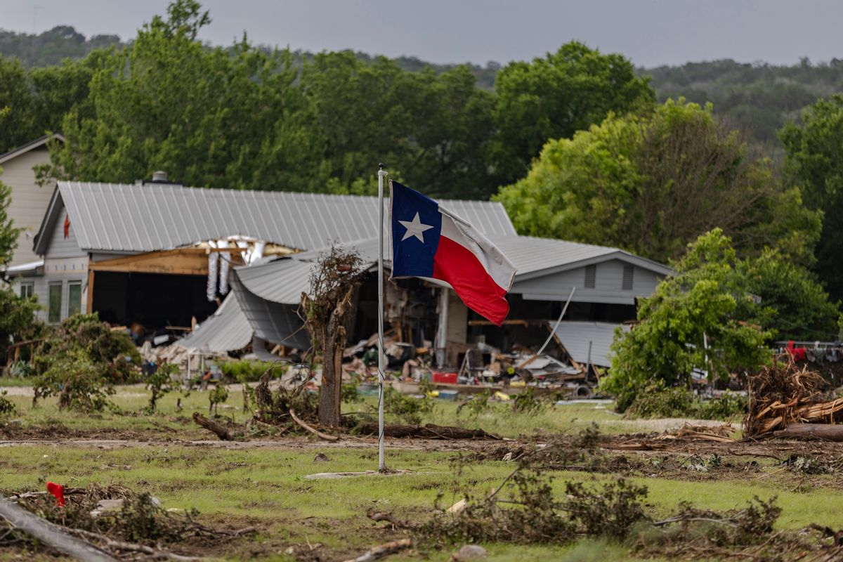 A blue, white and red striped flag with a single star — the Texas flag — in front of a partially destroyed house. 