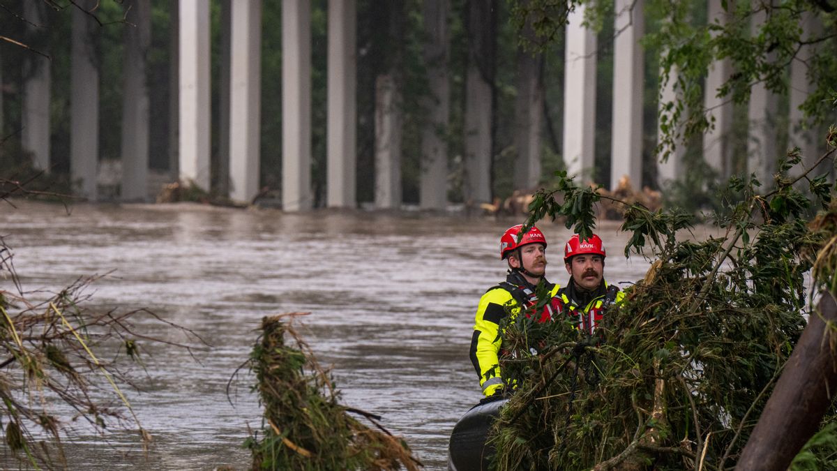 Two rescue workers in bright yellow and black gear with red helmets navigate floodwaters on an inflatable boat. They are surrounded by tree branches and debris, with a bridge supported by tall concrete columns in the background. 