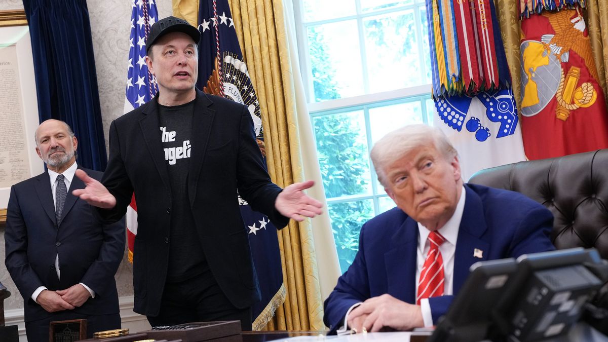 Elon Musk stands and gestures while speaking in the Oval Office, wearing a black jacket, black shirt and a cap. President Donald Trump sits at the Resolute Desk looking to his left toward Musk. Another man in a suit stands in the background. American and military flags and gold curtains are visible behind them.
