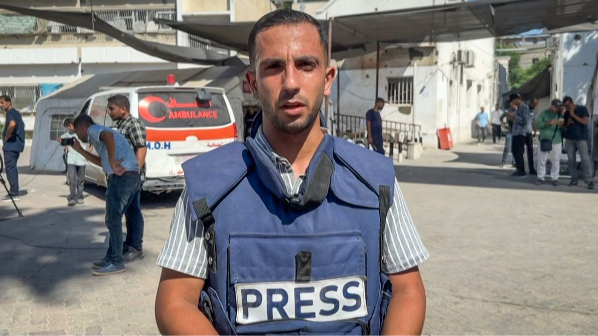 A male journalist wearing a blue press vest stands in the foreground, looking directly at the camera. Behind him, several people, including other journalists and bystanders, are gathered near an ambulance marked with "AMBULANCE" and "M.O.H" in both English and Arabic. The scene takes place outdoors in what appears to be a medical or emergency facility, with white buildings and some shade overhead. 