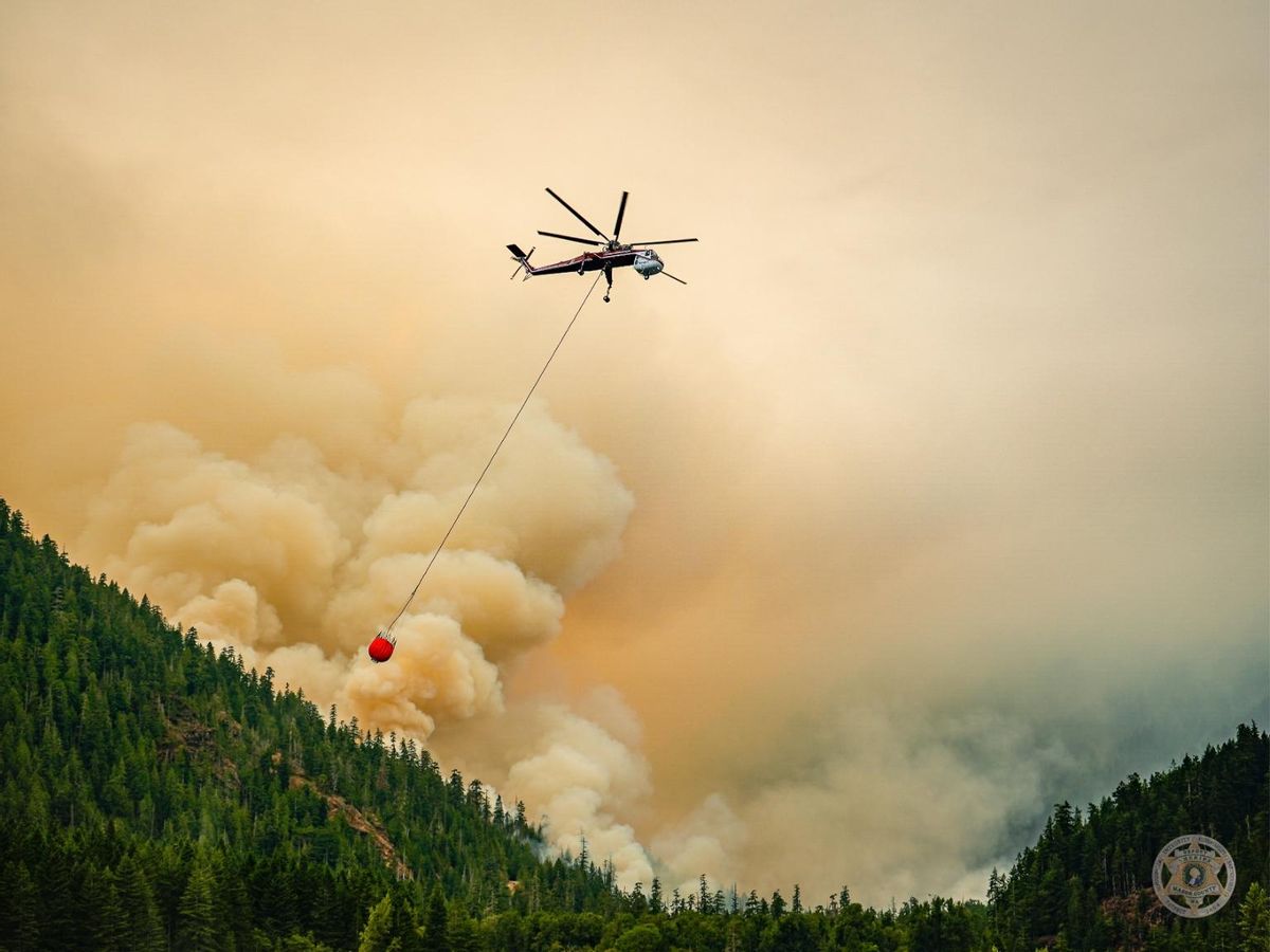 A helicopter with a bucket hanging from below flies over a smoky forest. 
