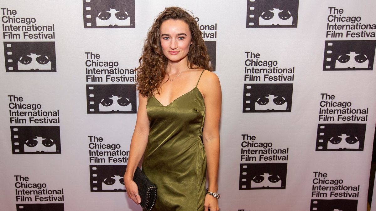 A woman with long, curly brown hair poses in front of a banner at the Chicago International Film Festival. She is wearing a sleeveless, olive green satin dress with thin straps and is holding a black clutch bag. The background features repeated logos of the festival, which include a stylized face peeking through film strips and the text "The Chicago International Film Festival."