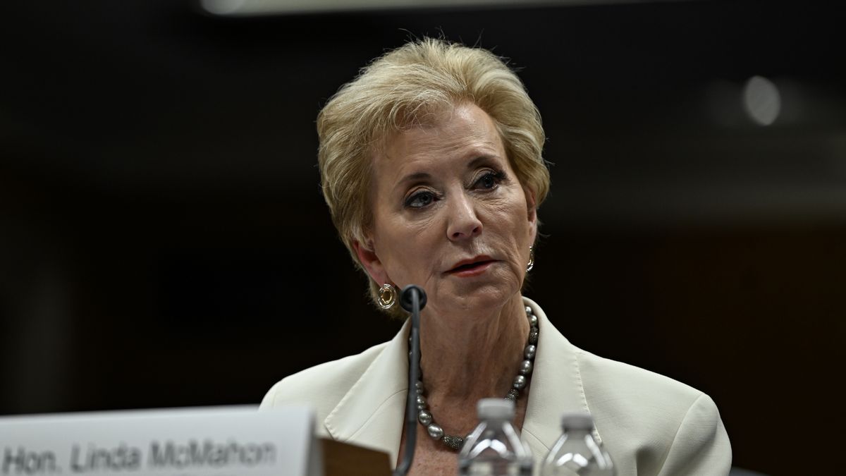A white woman with short blond hair wearing a white blazer and a pearl necklace sits at a table with a microphone in front of her. She appears to be speaking or listening intently during a formal event or hearing. A nameplate partially visible in the foreground reads "Hon. Linda McMahon." 