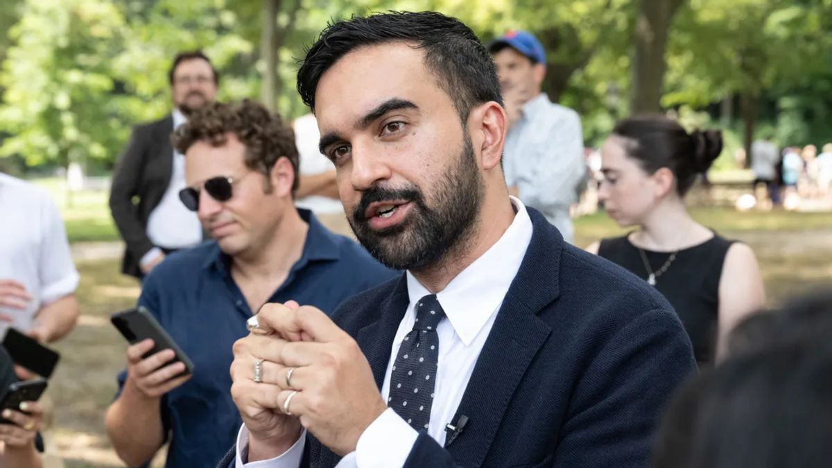 An Indian man wearing a dark blue suit stands looking to the left. People can be seen behind him.
