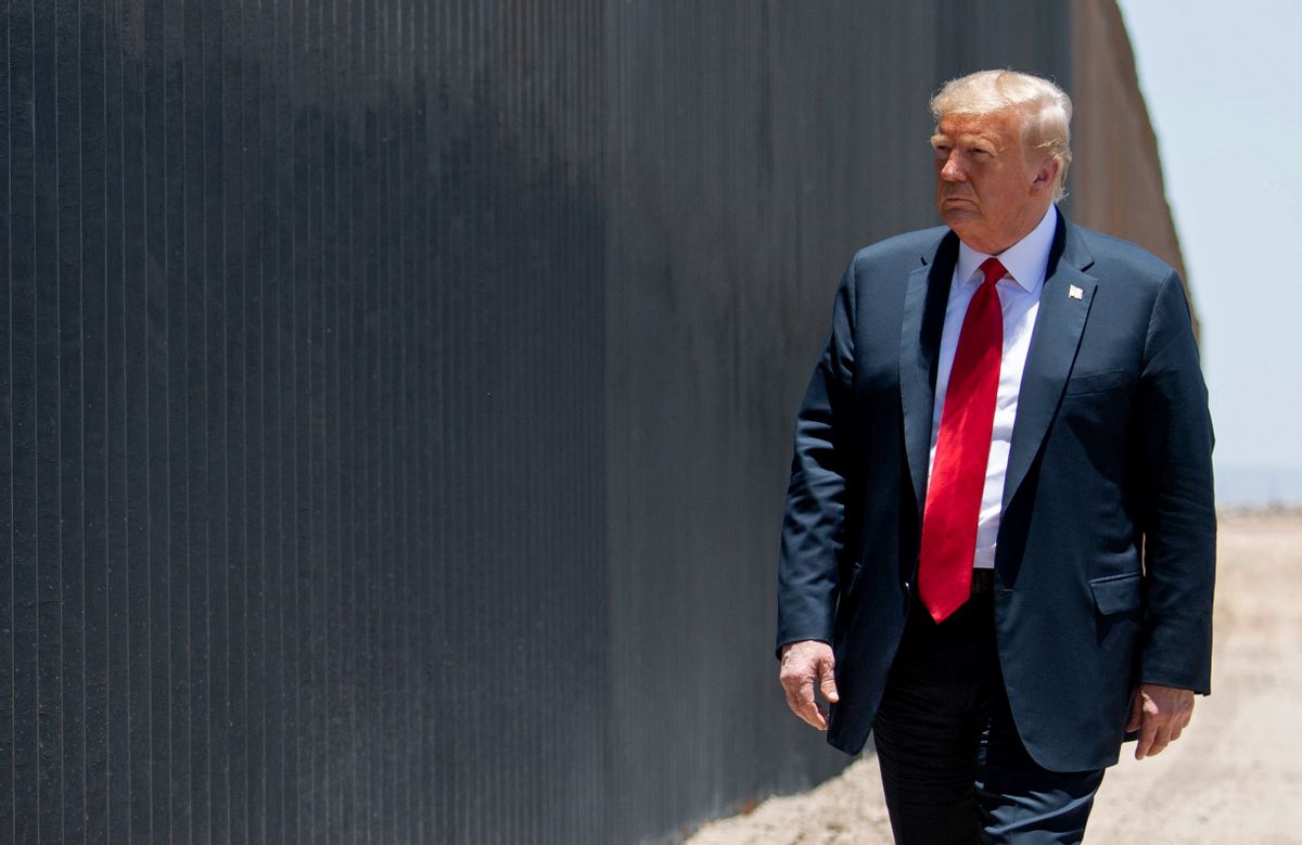 U.S. President Donald Trump, a white man in a red tie and dark suit, walks along a stretch of the U.S.-Mexico border wall, which looks like large metal slat fencing.