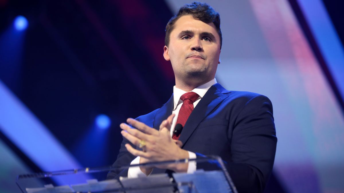 Charlie Kirk, a man in a navy blue suit with a red tie, stands behind a clear podium on a stage. He has a serious expression on his face. The background features blue and purple lights. 