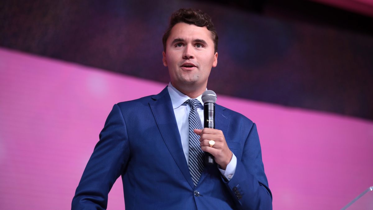 A man in a blue suit and striped tie speaks into a handheld microphone while standing on stage against a pink and purple background. He has short brown hair and is wearing a gold ring on his right hand.