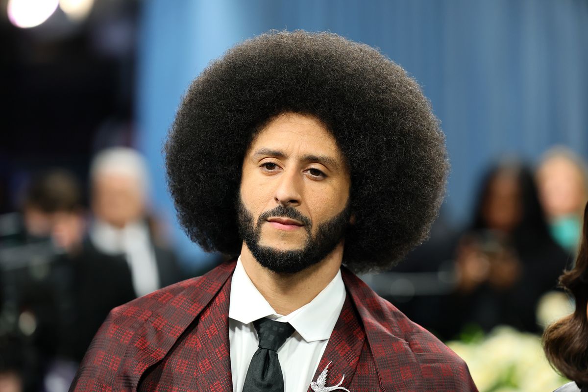 Colin Kaepernick, a Black man with a large afro and a trimmed beard, wearing a red and black patterned suit. 