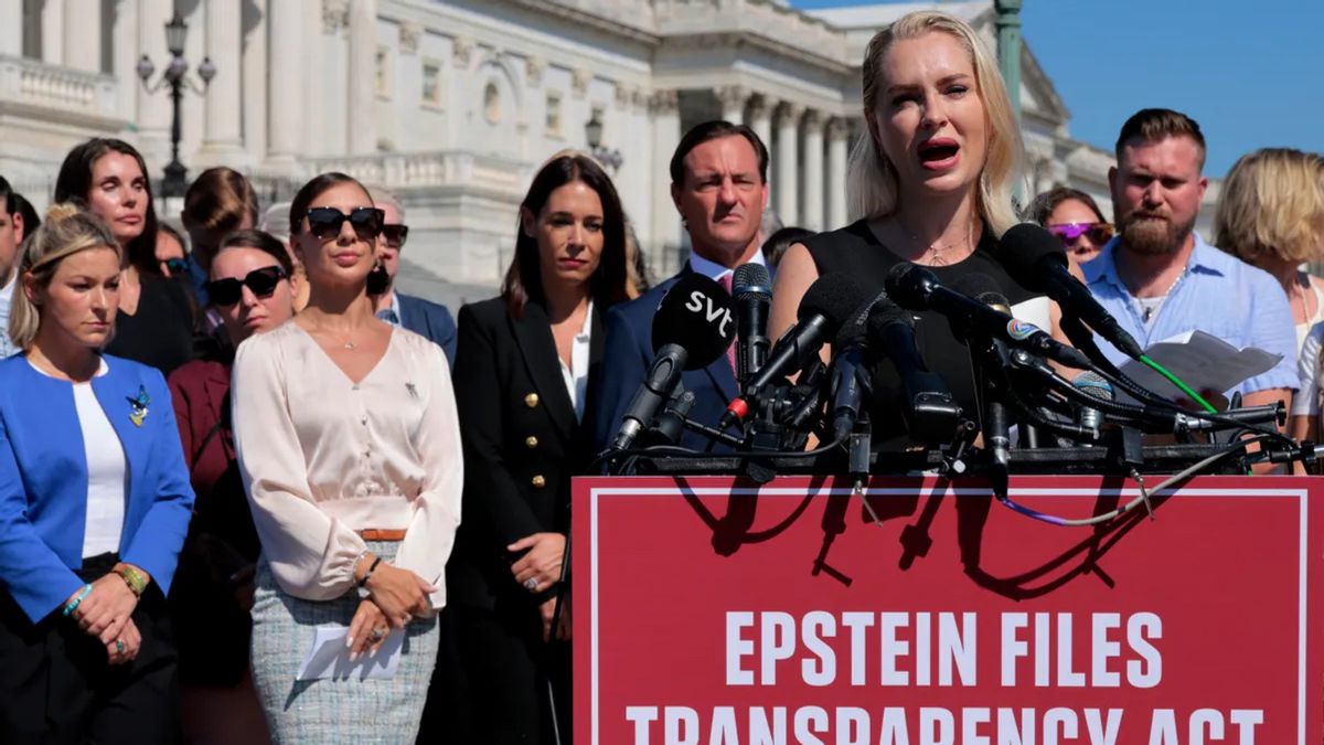A white woman speaks in front of a podium with a large red sign that says "EPSTEIN FILES TRANSPARENCY ACT." A group of people stand behind her and a U.S. Capitol building is visible in the background.
