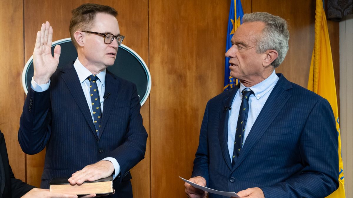 On the left, a middle-aged white man with short hair wearing glasses and a suit holds up one hand and keeps another on a book being held up by someone to the left of the camera. An older white man with short greying hair, also in a suit, speaks to the man on the left.