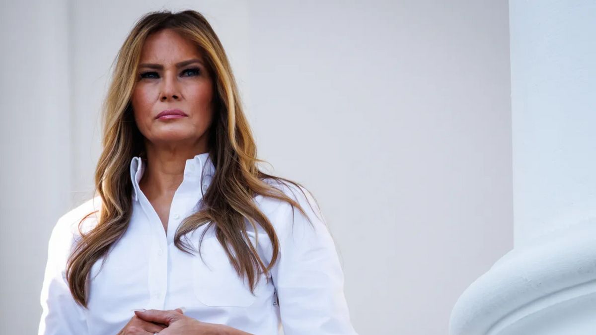 A white woman wearing a white button-down stands with her hands clasped.