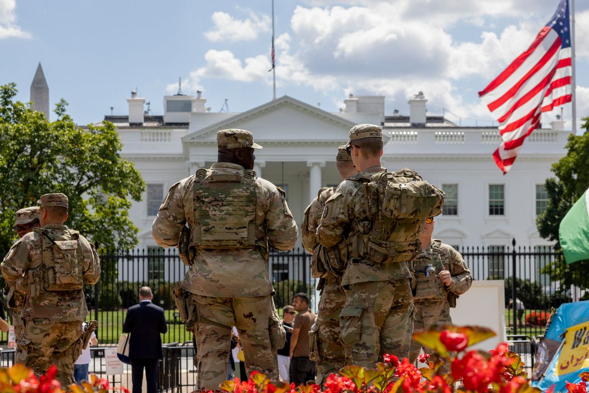 A group of National Guardsmen in camo military wear standing outside of the White House. A U.S. flag is on the right. 