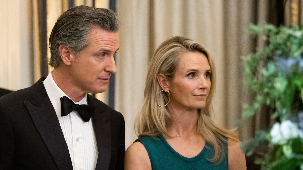 A white man with salt and pepper hair in a tuxedo and a blonde white woman wearing gold hoops and a green sleeveless dress look off to the right of the camera.