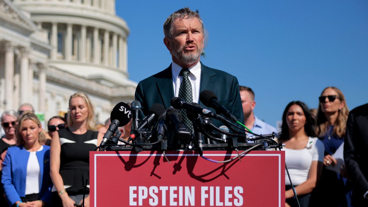  A white man with short greying hair and a beard wearing a suit stands in front of a podium with a large red sign that reads, "EPSTEIN FILES TRANSPARENCY ACT." A large group of people stand behind him. They are in front of the U.S. Capitol, a white dome building.