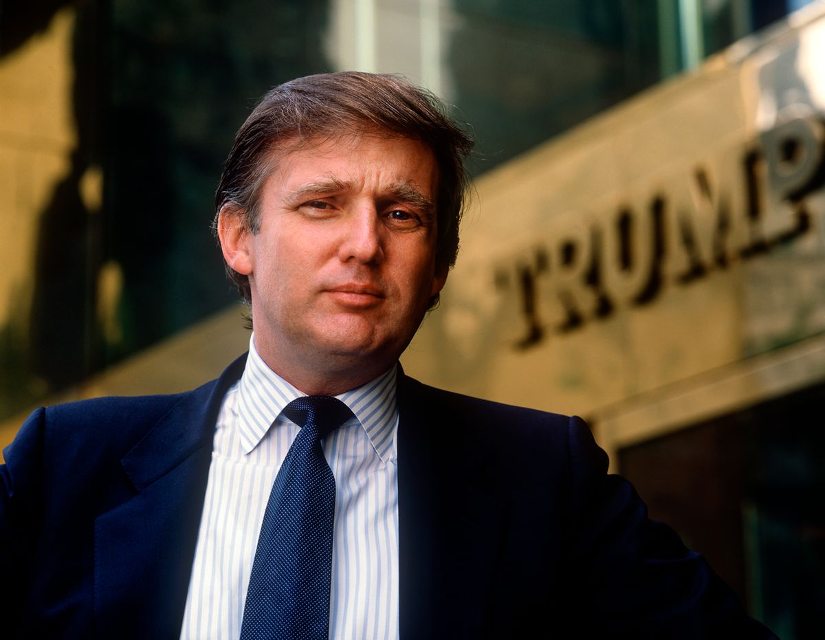 A young Donald Trump — a white man with combed over blonde hair wearing a suit — standing in front of a building labeled "TRUMP." 