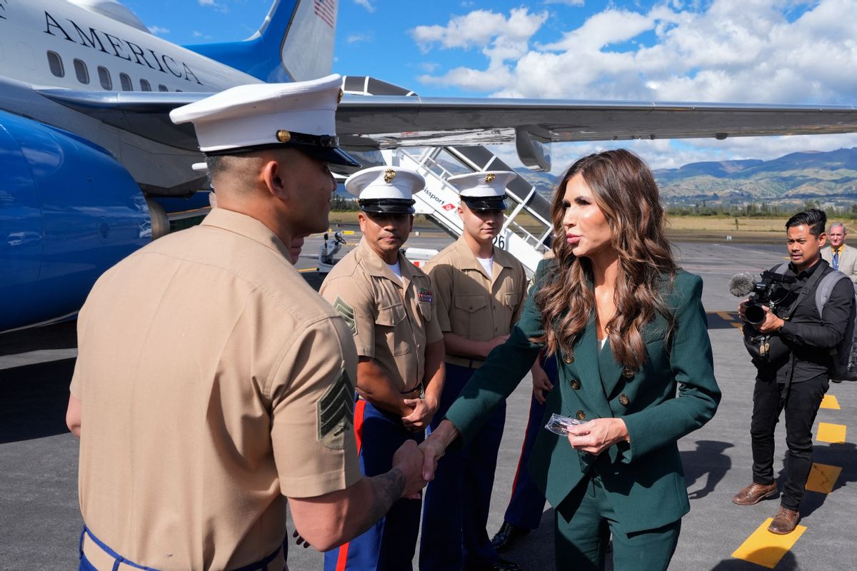 A white woman with long wavy brunette hair wearing a dark green suit (right) shakes hands with uniformed men (left) in front of a plane. 