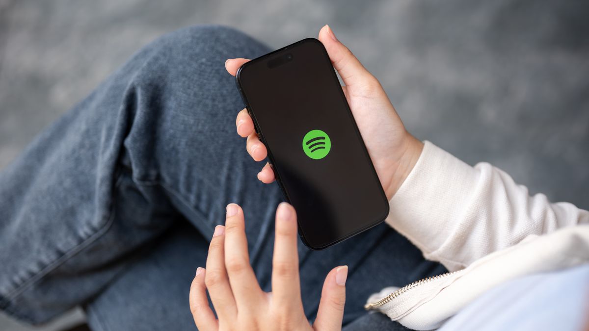 Person wearing jeans and white sweatshirt is shown holding a black cellphone displaying the green Spotify logo