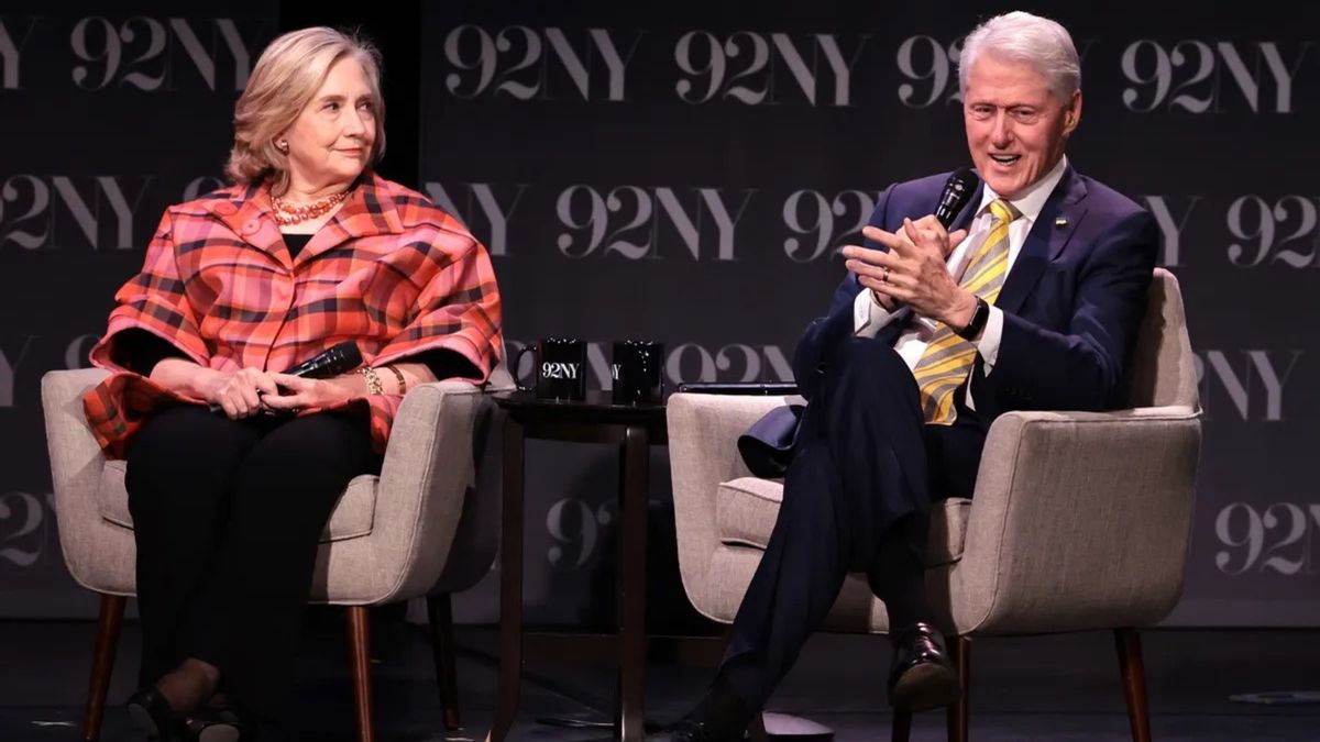 Hillary Clinton, a white woman sitting down, looks at her husband Bill Clinton, a white man also sitting down, as he speaks into a microphone.