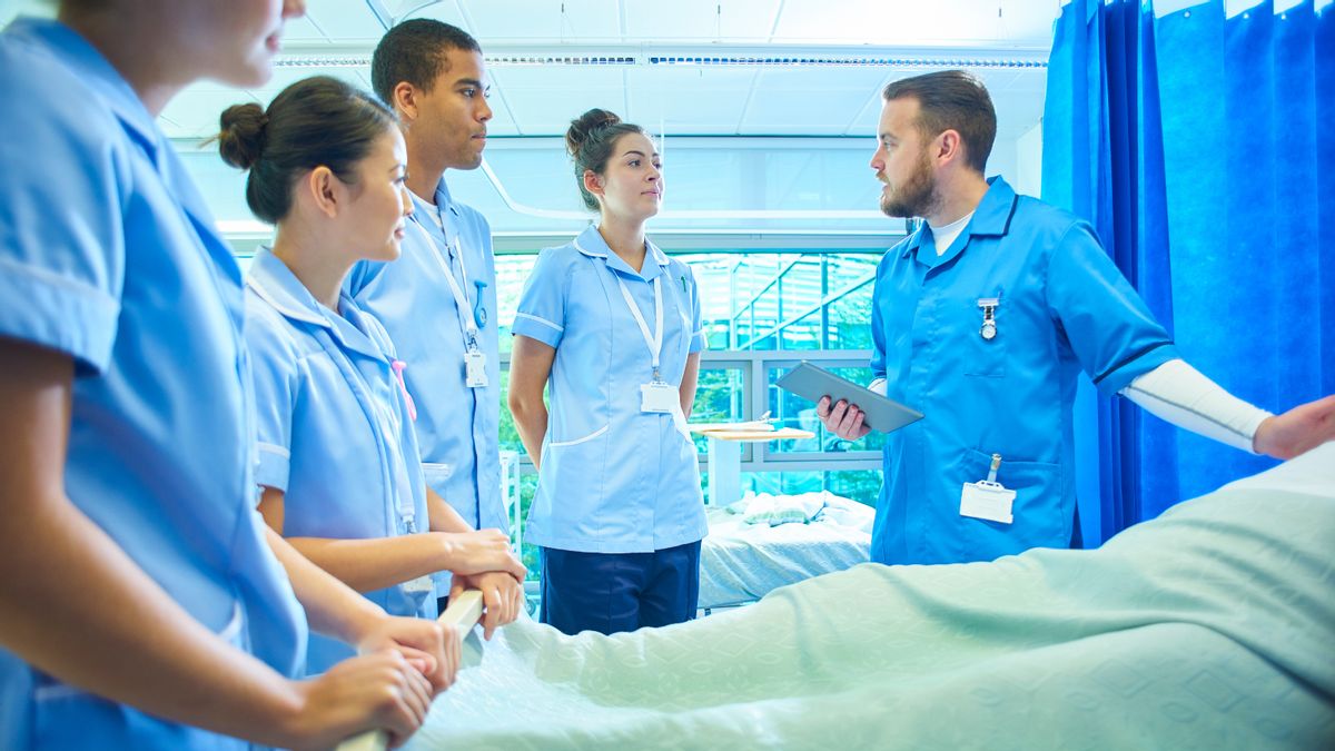 Four students in blue scrubs stand next to a hospital bed and listen to an instructor holding a clipboard who is also wearing blue scrubs. 