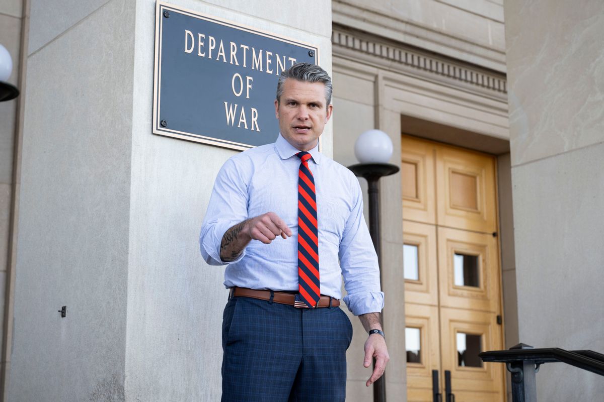 Defense Secretary Pete Hegseth, a white man with salt and pepper hair wearing a blue and red tie and American flag belt buckle, stands in front of a sign that says, "DEPARTMENT OF WAR."