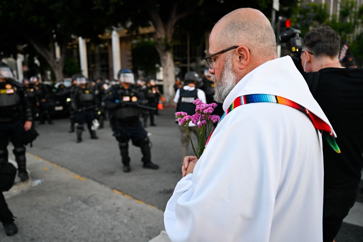 A priest wearing white robes with a colorful stole holds a bunch of purple flowers. He is standing with his head bowed in front of law enforcement wearing heavy tactical gear. 