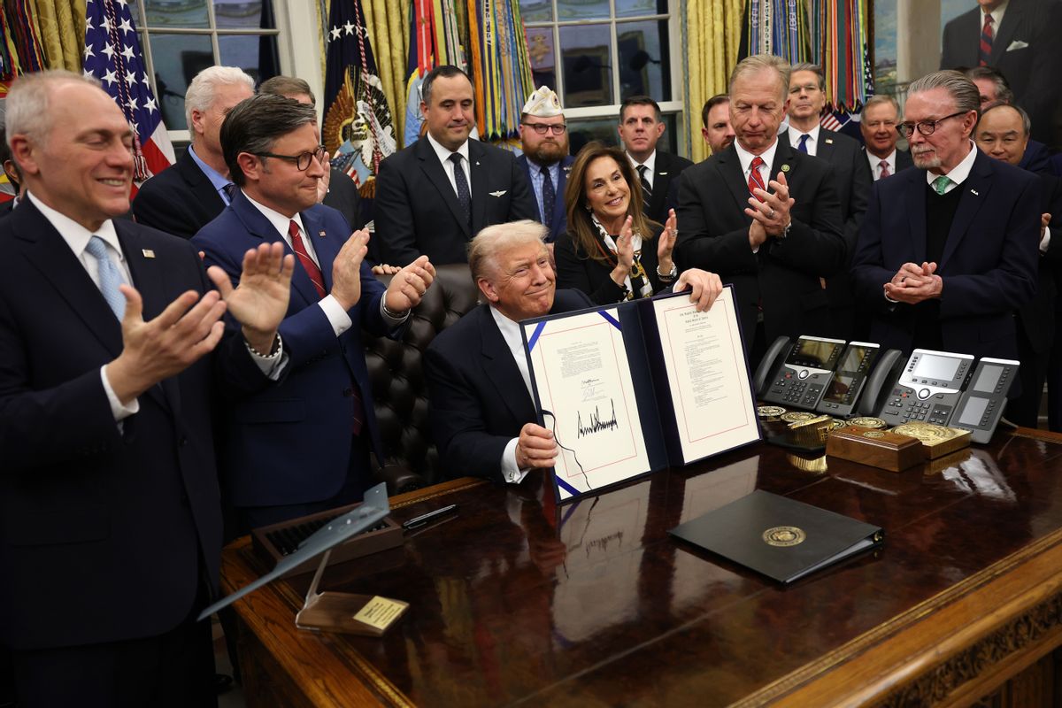 U.S. President Donald Trump holds up a signed bill and smiles sitting behind a desk in the Oval Office. People surrounding him clap. 