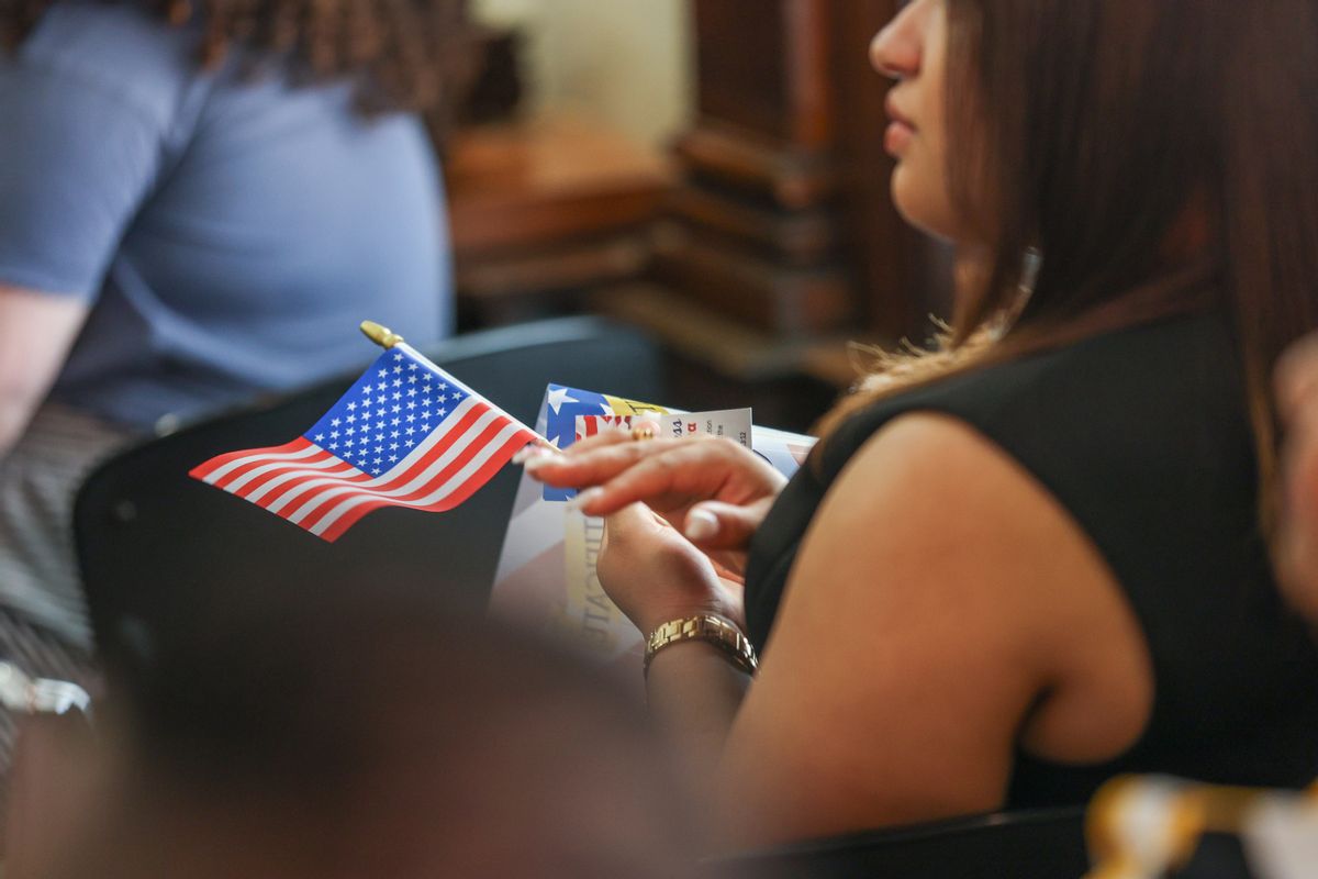 A woman seated in a chair holds a U.S. flag between her hands and claps at a citizenship ceremony. 