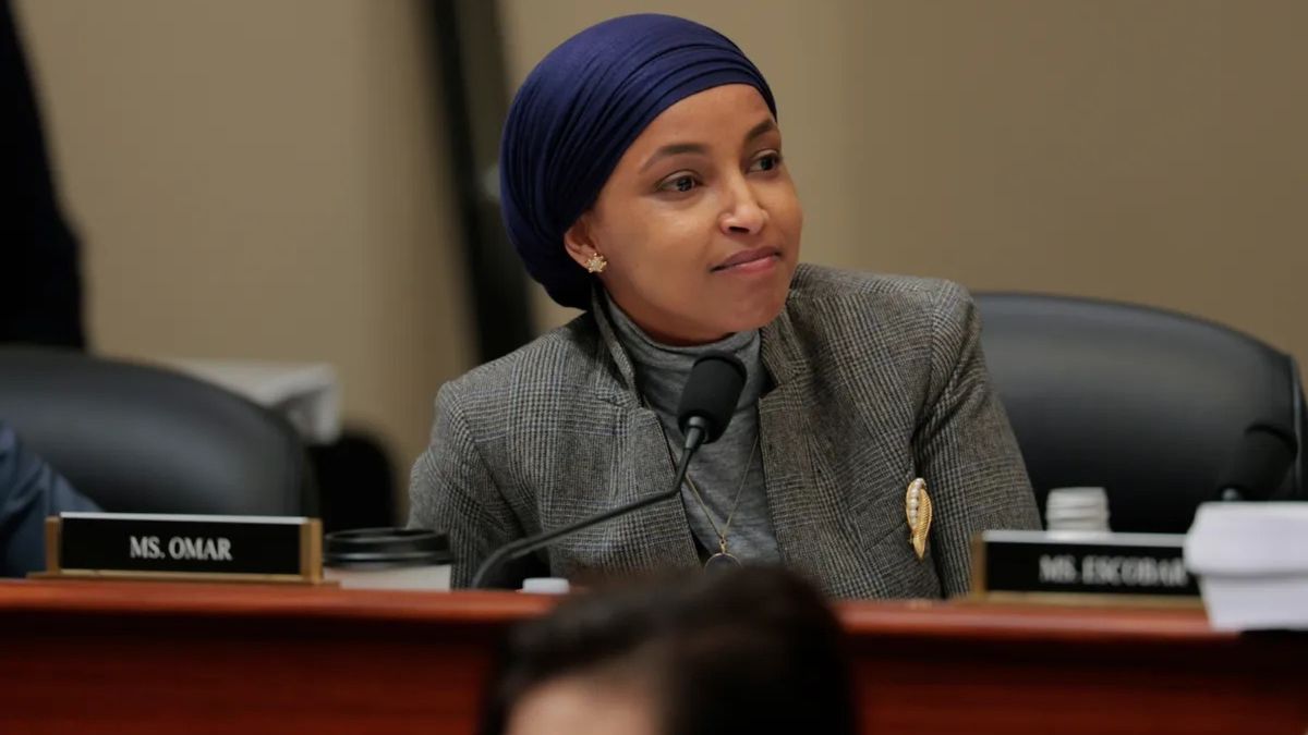 Ilhan Omar, a Black woman wearing a gray suit jacket and dark blue headscarf, is photographed looking towards the right. She is sitting at a desk with a name tag that says "MS. OMAR" in front of her.