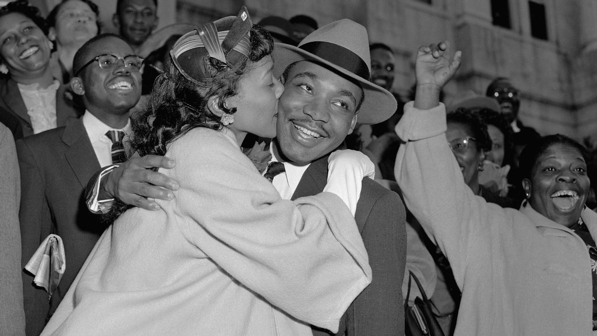 A black and white image shows a Black man being kissed on his left cheek by a Balck woman. A crowd of people, many of them Black, can be seen happy and smiling behind him.