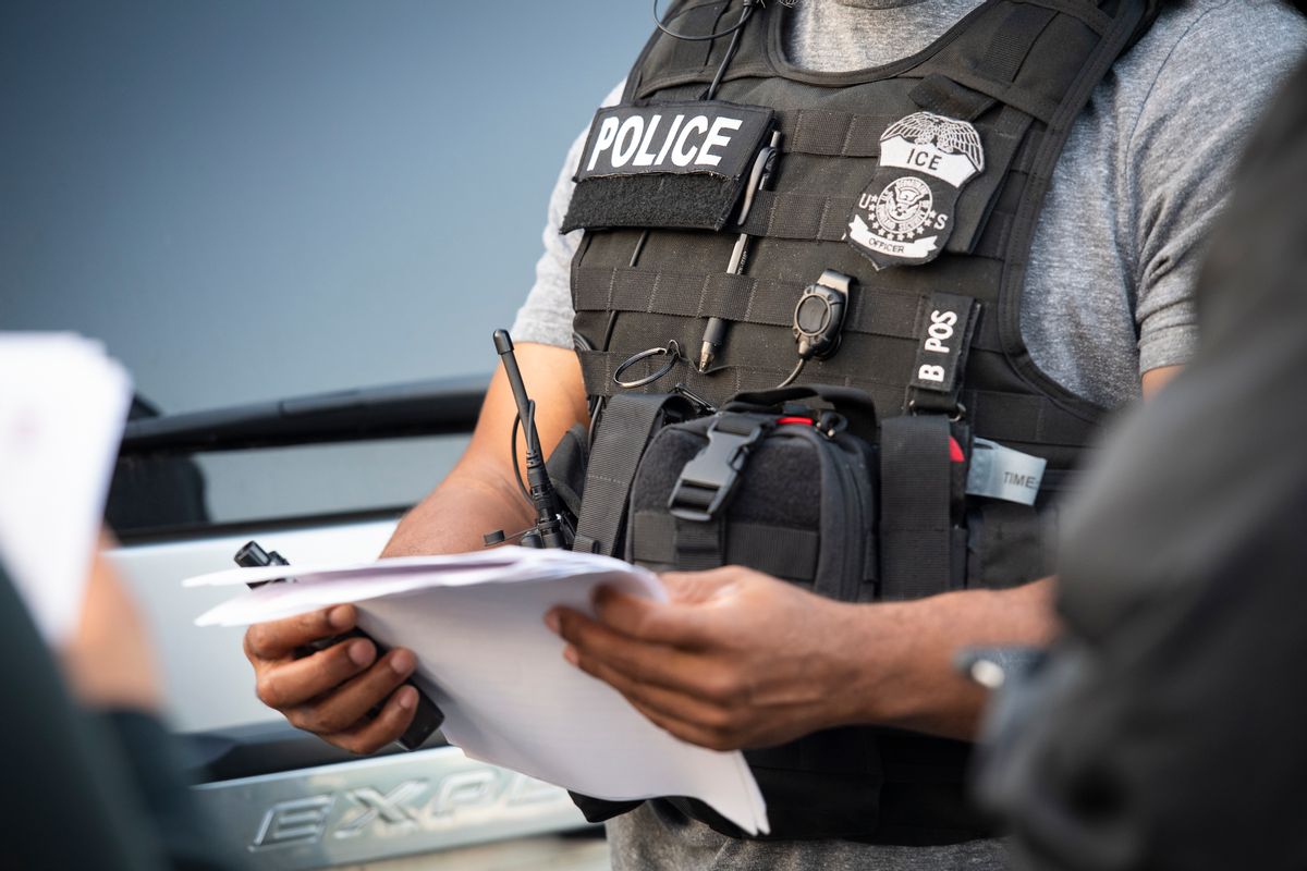 An ICE officer wearing a badge and vest holds a piece of paper. 