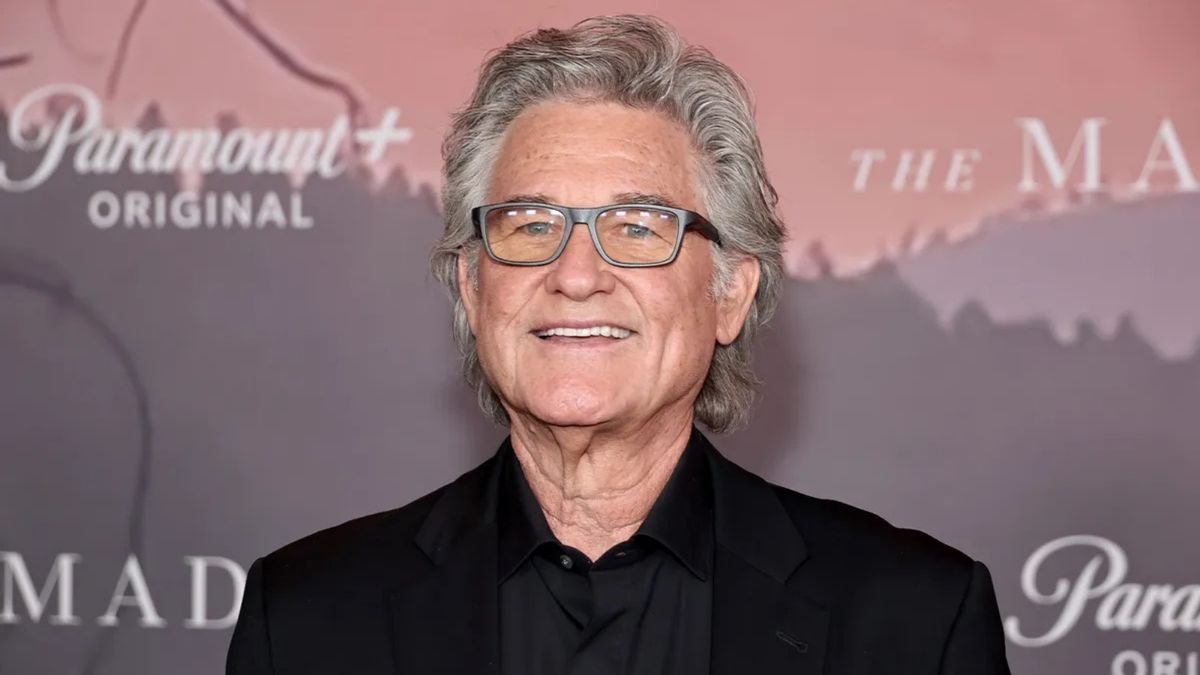 The actor Kurt Russell, an older white man with medium length gray hair and glasses, smiling.