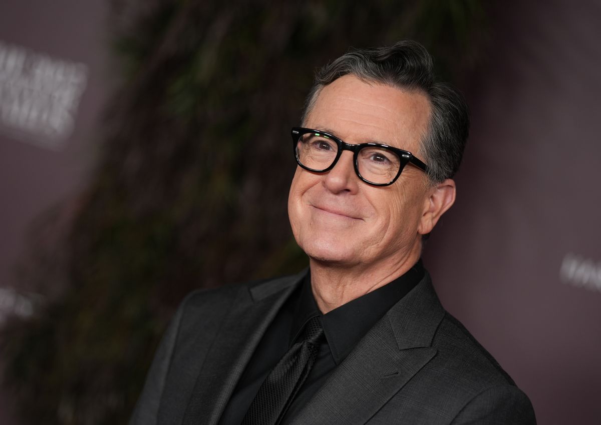 Stephen Colbert, a white man with dark, slicked back hair and glasses, wearing an all-black suit.