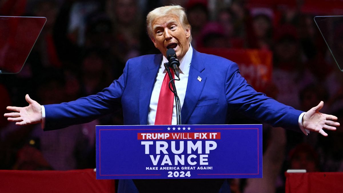 U.S. President Donald Trump stands in front of a podium with his arms spread and a large audience behind him. 