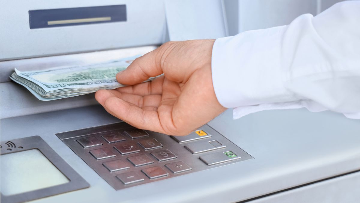 A close-up shot of an ATM with a hand holding a stack of $100 bills. 