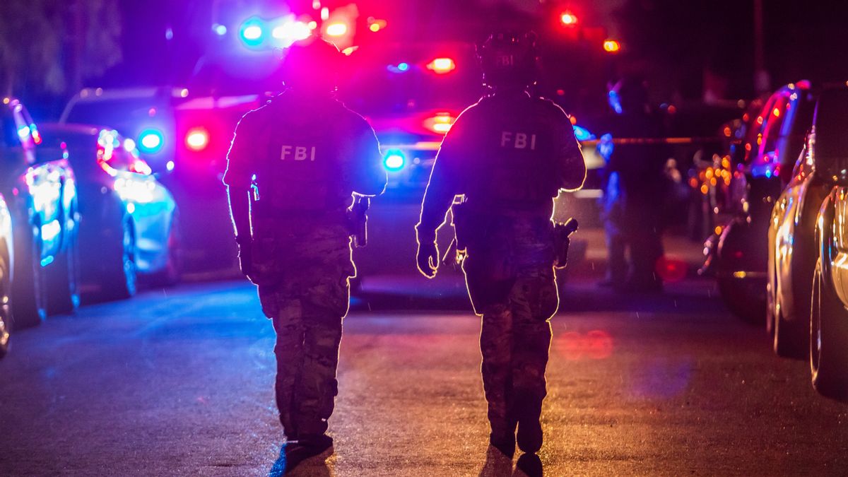 Two FBI agents backlit by red and blue police lights walking down a road at night.