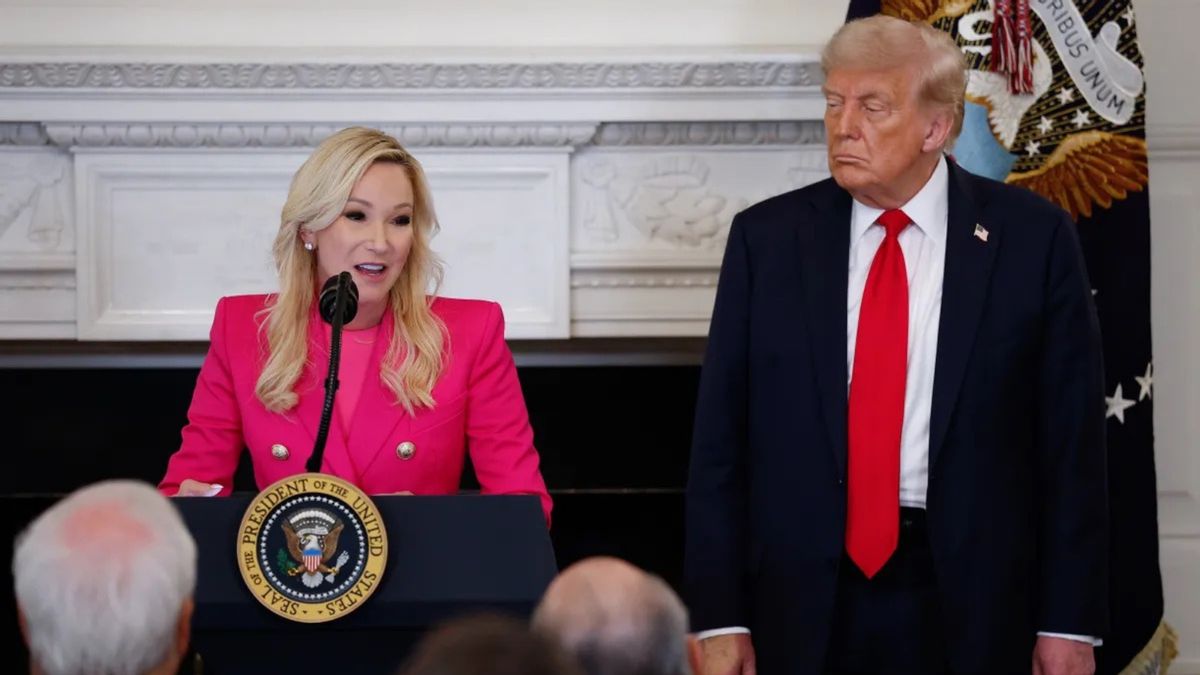 Paula White-Cain, a middle-aged white woman with platinum blonde hair, speaks at a podium wearing a neon pink suit. Donald Trump stands to her left, watching.