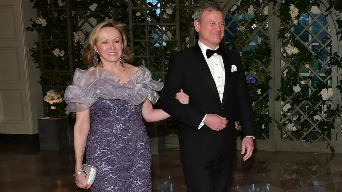 An older white woman with shoulder length blonde hair wearing a purple dress and an older white man wearing a black tuxedo. 