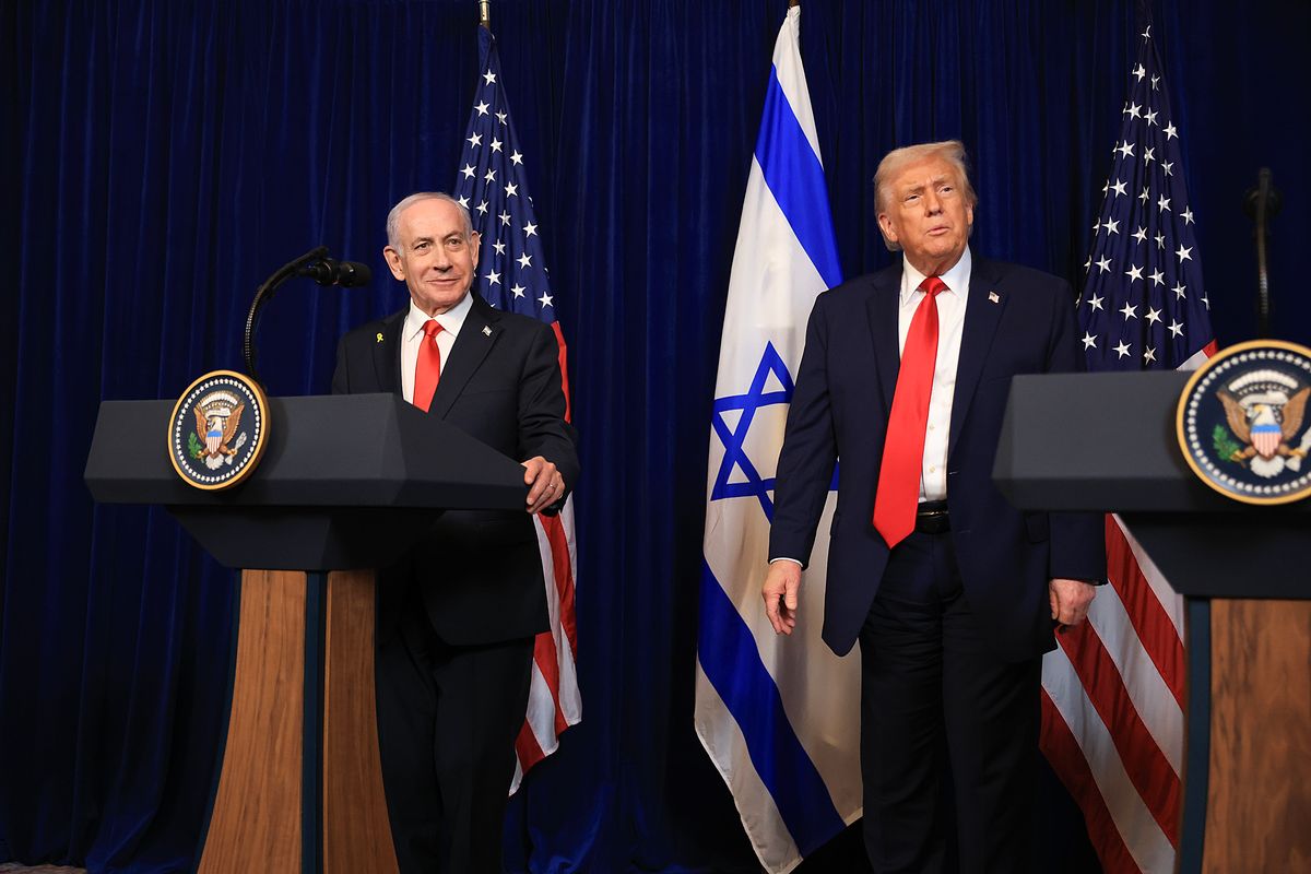 Two older white men stand at podiums. Netanyahu, with short cropped white hair, stands on the left, while Trump, with longer blond hair, stands on the right. US and Israel flags are on poles behind them. 