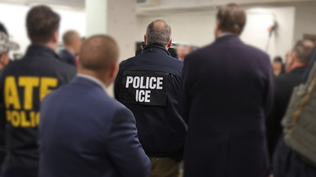 A federal agent stands facing away from the camera wearing a jacket that reads "ICE."