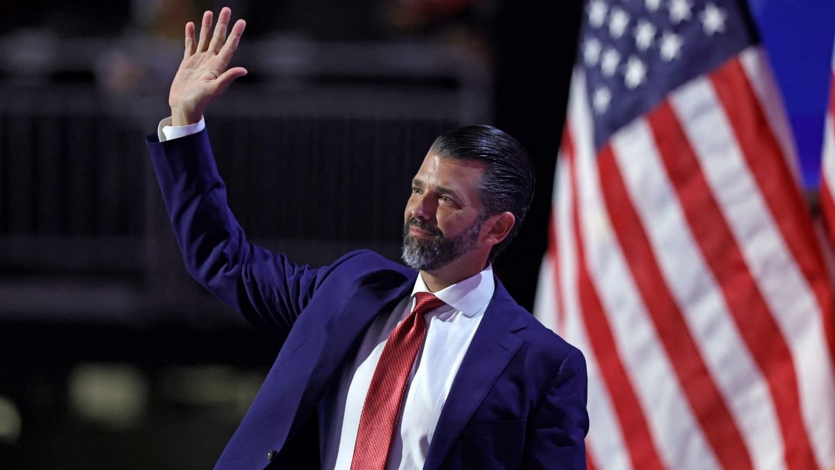 Donald Trump Jr., a white man, wears a suit and waves while standing in front of an American flag.